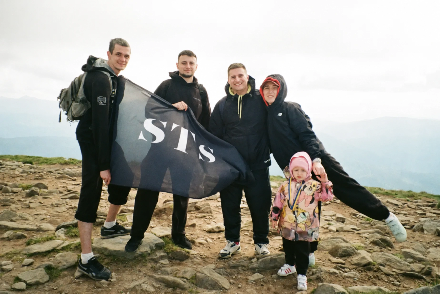 Five people, including a small child, proudly hold a flag on a rocky mountain summit.